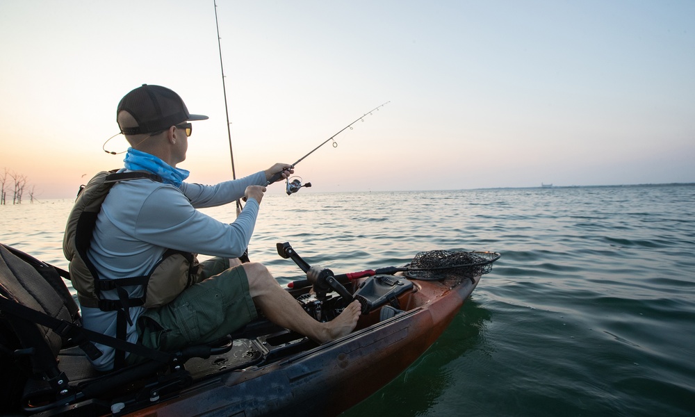 A man in a baseball cap fishes from a kayak on calm ocean water with a fishing rod extended toward the horizon.