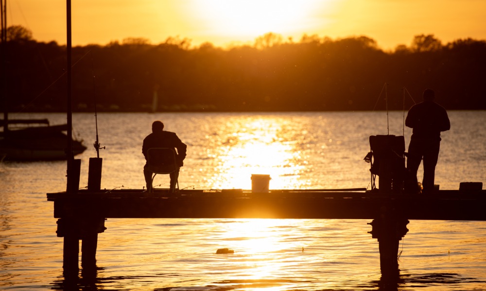 The silhoutte of two people standing on a dock and fishing during sunset, with the sun low in the sky.