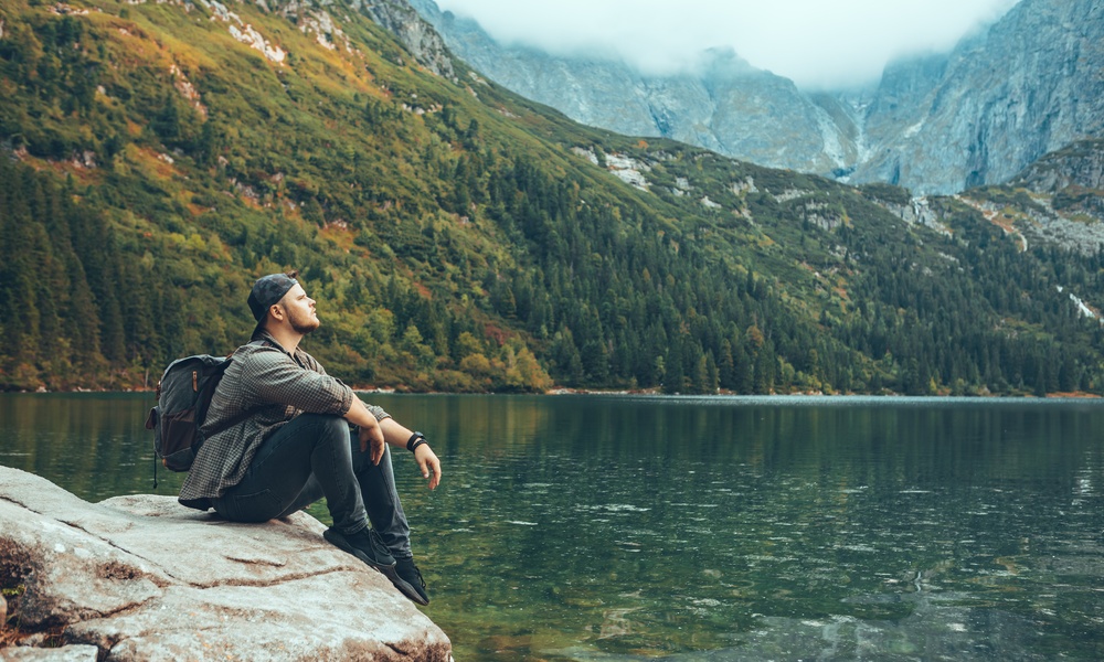A man wearing a backpack sits on a rock by a clear mountain lake and looks across the lake toward the mountains.