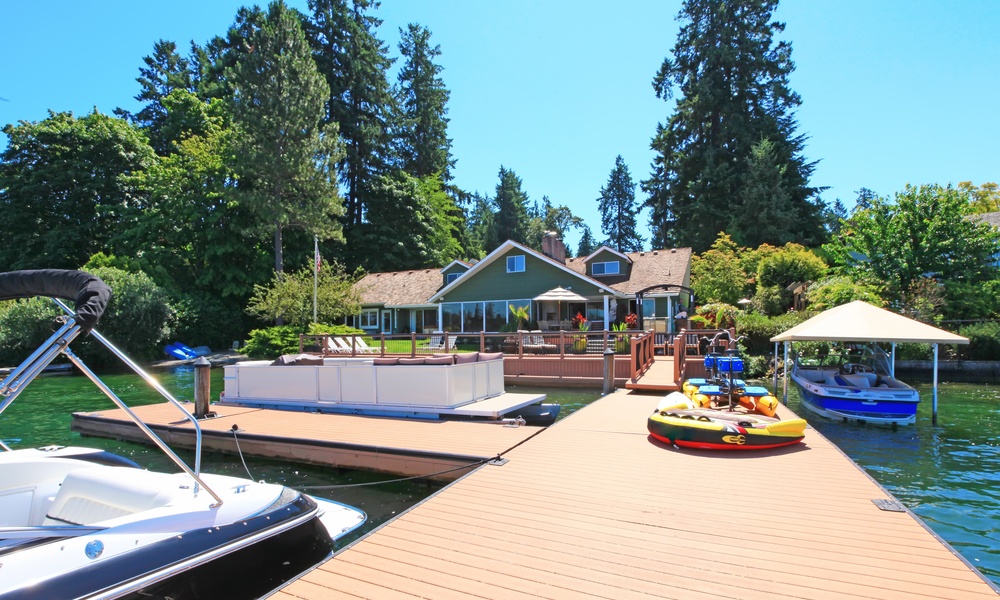 Lakeside home with a large wooden dock, multiple boats, and water gear, surrounded by tall trees under a clear blue sky.