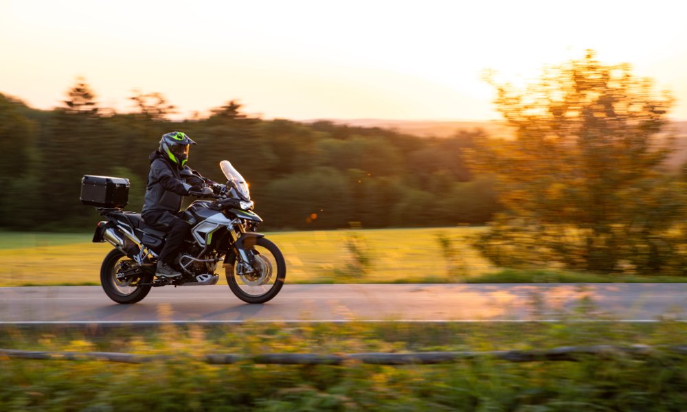 Motorcyclist rides touring bike along rural road at sunset, with trees and fields blurred in warm evening light.