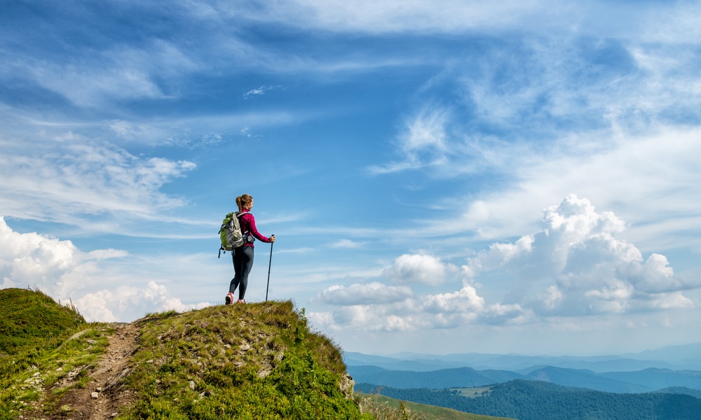 A young woman with a walking stick standing on a mountain looking out at the view. She also has a backpack on.