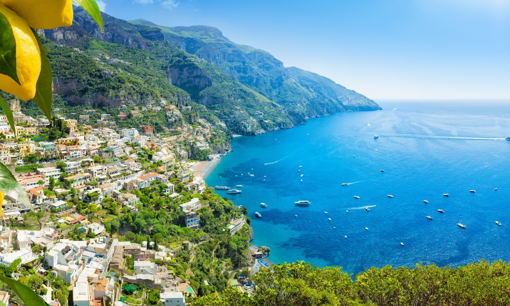 A landscape view of Italy, overlooking a coastal town with an up-close view of a lemon tree. Boats are in the water.