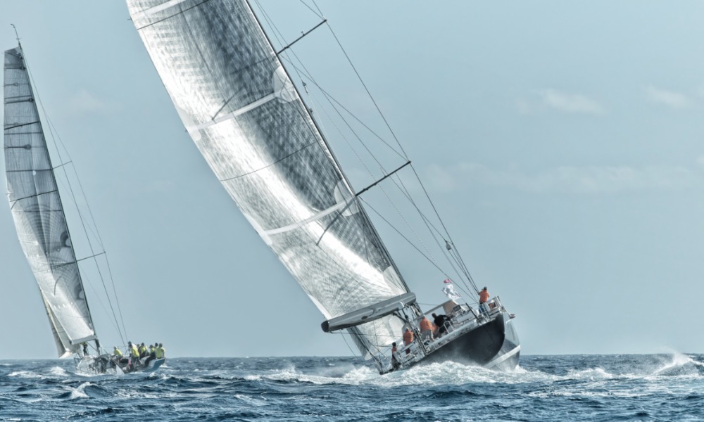 Two sailboats race on choppy water under an overcast sky, sails taut as the lead boat heels sharply.