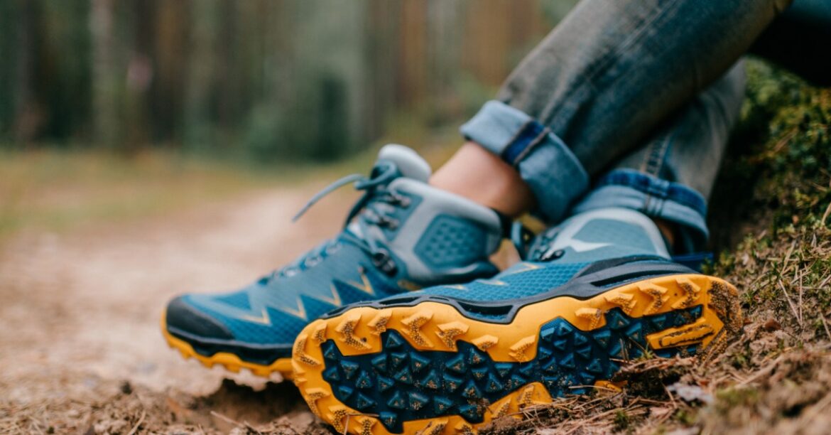 A person wearing blue hiking shoes and rolled jeans resting their feet on a dirt path beside mossy ground.